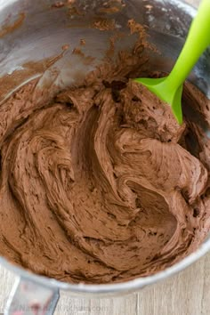 a metal bowl filled with chocolate frosting on top of a wooden table next to a green spatula