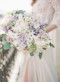 a woman holding a bouquet of white and purple flowers on her wedding day in front of a fence