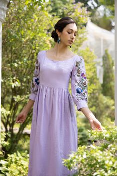 a woman in a purple dress is standing near some bushes and trees with her hands out to the side