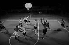 black and white photograph of men playing basketball on court at night with lights from overhead