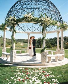 a bride and groom standing under a gazebo with flowers on the ground in front of them