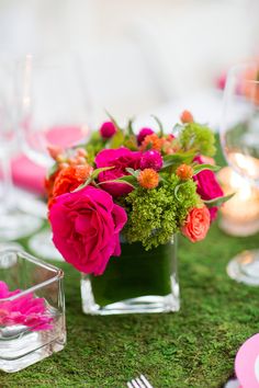 a vase filled with pink flowers on top of a green moss covered tablecloth and place settings