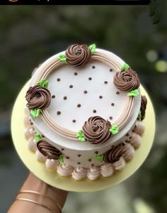 a hand holding a cake with chocolate icing and green leaves on it, in front of a blurry background