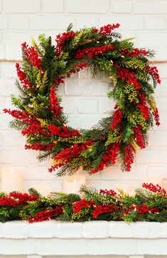 a christmas wreath with red berries and greenery hanging on a brick wall next to candles