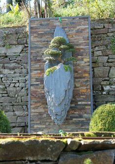 a stone sculpture with trees on it in front of a rock wall and shrubbery