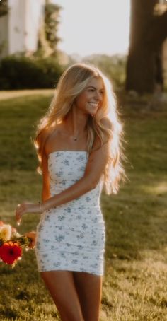 a woman in a short white dress holding flowers and smiling at the camera while standing on grass