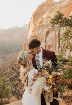 a bride and groom kissing in front of the mountains at their wedding day, with an orange flower bouquet