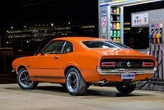 an orange muscle car parked at a gas station in front of a fuel pump with nozzles on it