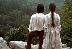 two people standing on top of a mountain looking out at the trees and hills in the distance