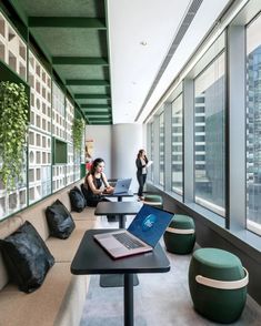 two women sitting at tables with laptops in an office setting, overlooking the city