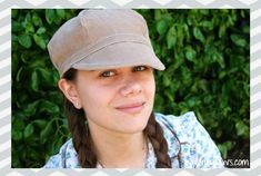 a woman with braids wearing a hat and looking at the camera while smiling in front of some bushes