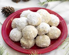 a red plate filled with snowball cookies on top of a white table cloth next to pine cones