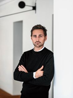 a man standing next to a white wall with his arms crossed and looking at the camera