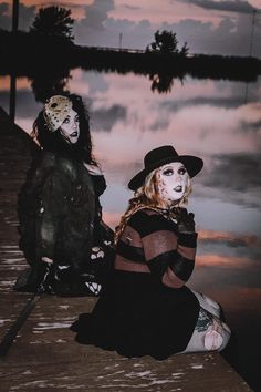 two women dressed in costumes sitting next to each other on a dock near water at sunset