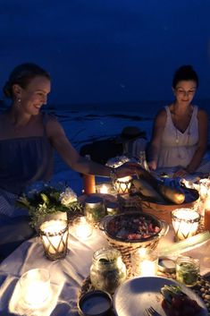 three women sitting around a table with food and candles on it at night, near the ocean