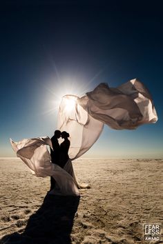 two people are standing in the sand with their arms around each other and one person is holding a white fabric