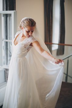 a woman in a wedding dress standing by a window with her veil flying over her head