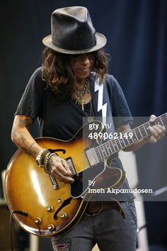 a man with long hair playing an electric guitar at a music festival, wearing a fedora and black t - shirt