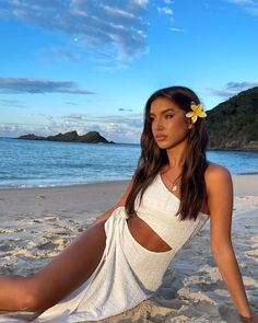 a beautiful young woman sitting on top of a sandy beach next to the ocean with a flower in her hair