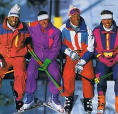 four skiers sitting on a ski lift posing for a photo in their colorful outfits