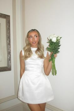 a woman in a short white dress holding a bouquet of flowers next to a mirror