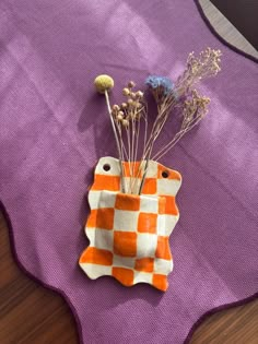 an orange and white checkered vase sitting on top of a purple table cloth next to dried flowers