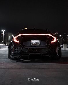 the rear end of a black sports car at night with city lights in the background