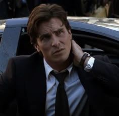 a man in a suit and tie leaning against a car with his hand on his ear