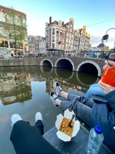 two people sitting on the edge of a river with food in front of them and drinking water