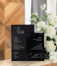 two black menu cards sitting on top of a table with white flowers in the background