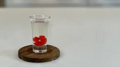 a small glass filled with liquid and some red flowers on top of it, sitting on a wooden coaster