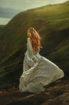 a woman in a white dress sitting on top of a hill with her hair blowing in the wind