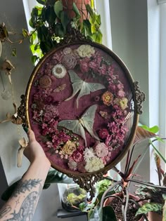 a person holding up a painting with flowers on it in front of a window sill