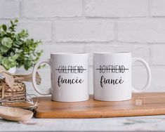 two white coffee mugs sitting on top of a wooden table next to a potted plant