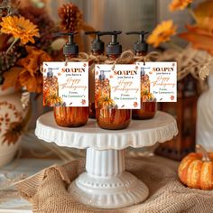 three bottles of soap sitting on top of a white cake plate with pumpkins in the background