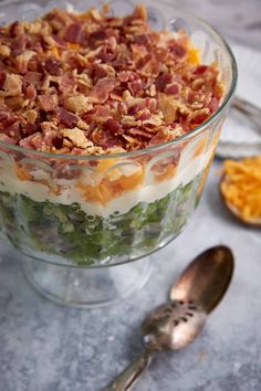 a layered salad in a glass bowl with bacon on top and other food items around it