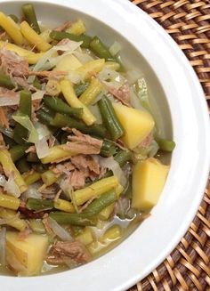 a white bowl filled with green beans and meat next to a brown woven place mat