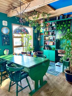 a green table and chairs in a room with bookshelves, potted plants and windows