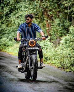 a man riding on the back of a motorcycle down a road next to lush green trees