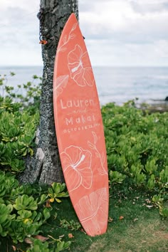 an orange surfboard leaning against a tree on the grass by the ocean with flowers painted on it