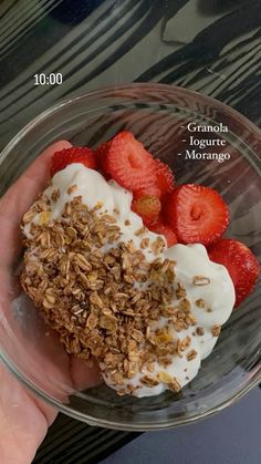 a hand holding a glass bowl filled with granola and yogurt next to strawberries