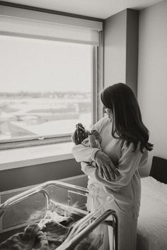 a woman holding a baby in her arms while standing next to a hospital bed and looking out the window