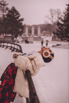 a teddy bear wearing a hat and scarf leaning on a rail in the snow near a park