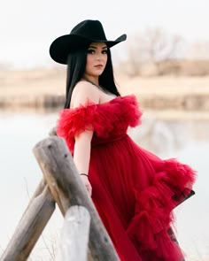 a woman with long black hair wearing a red dress and cowboy hat, leaning on a fence