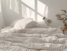 an unmade bed with white sheets and pillows in front of a window, next to a potted plant