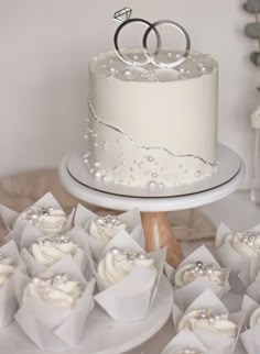 two wedding rings on top of a white cake surrounded by cupcakes and napkins