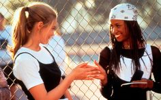 two young women standing next to each other near a chain link fence, talking and laughing