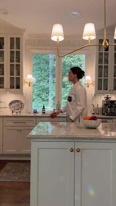 a woman standing at the counter in a kitchen with white cabinets and marble counter tops