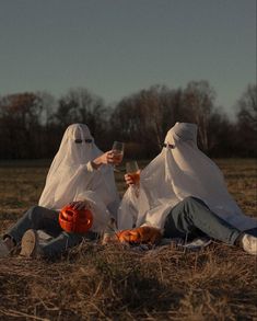 two people dressed in white sitting on the ground holding wine glasses and pumpkins,