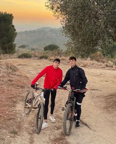 two men standing next to each other on dirt road with bikes in front of them